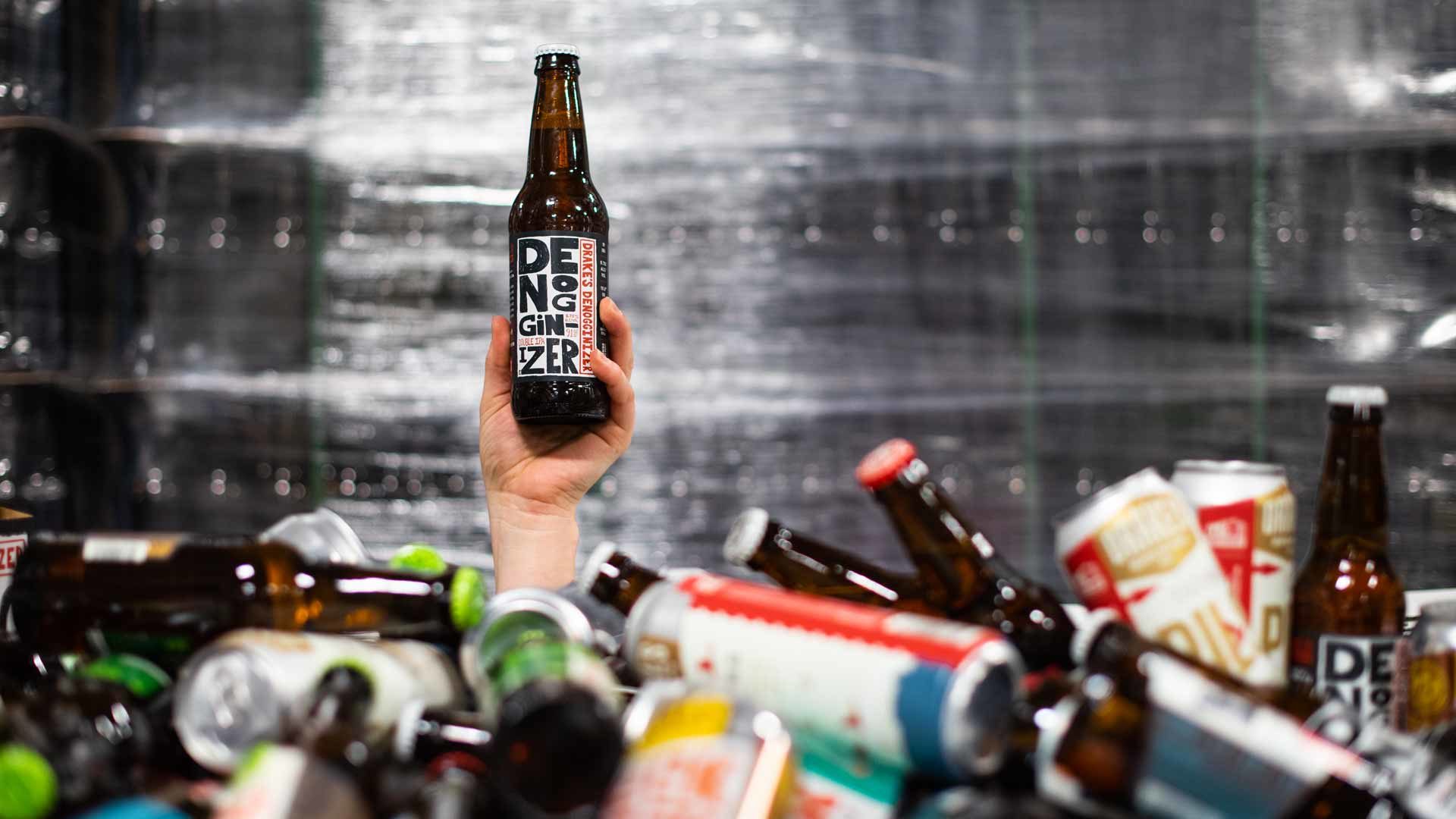 A bottle of Denogginizer Double IPA being held above a pile of cans and bottles