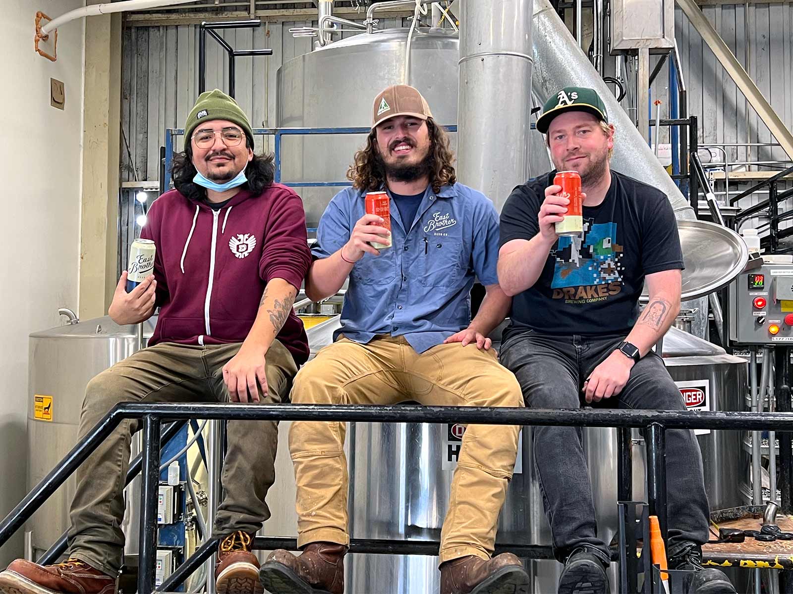 Three men in a brewery hold up cans of craft beer, smiling