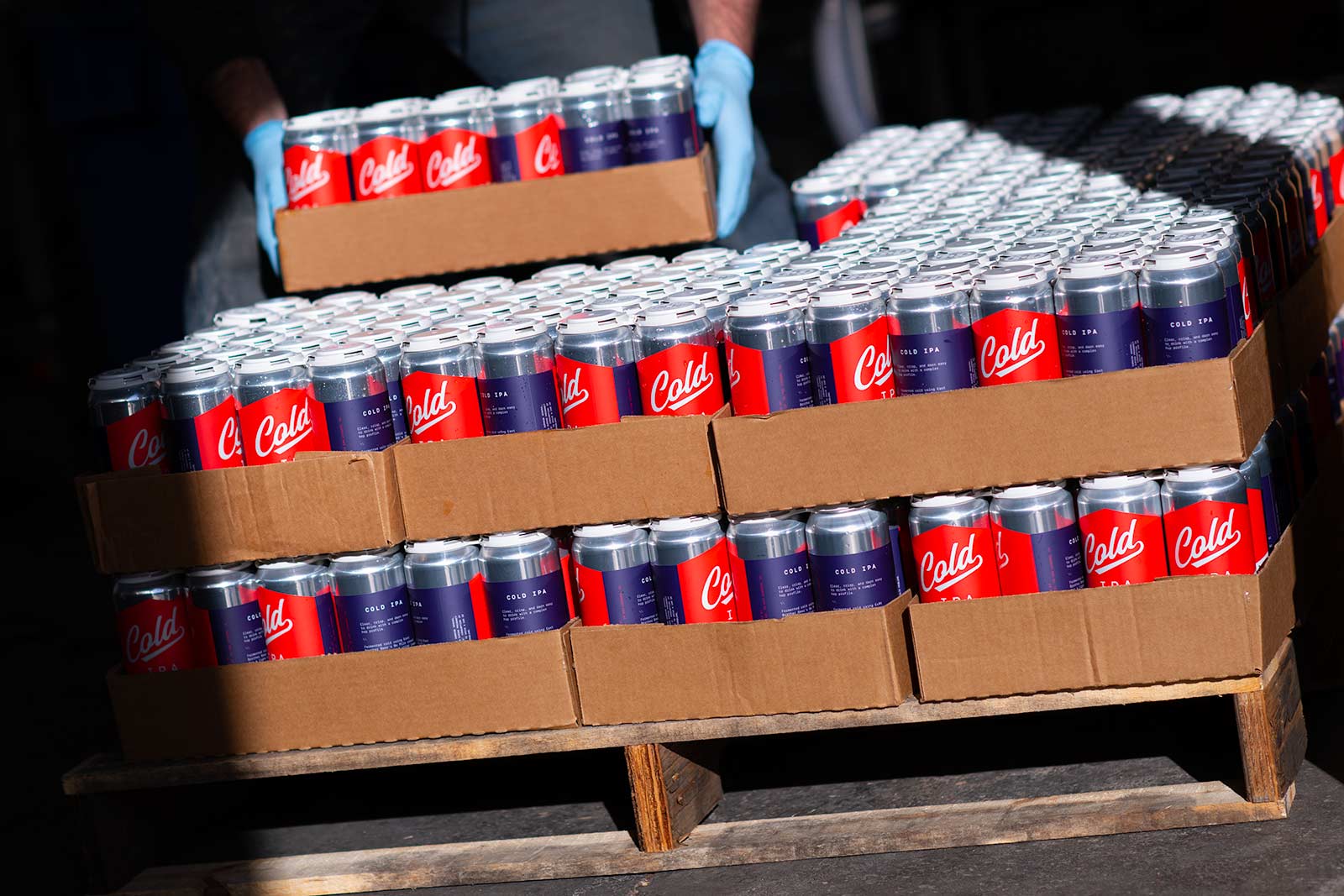Hands in blue gloves stacking cases of canned beverages labeled "Cold”