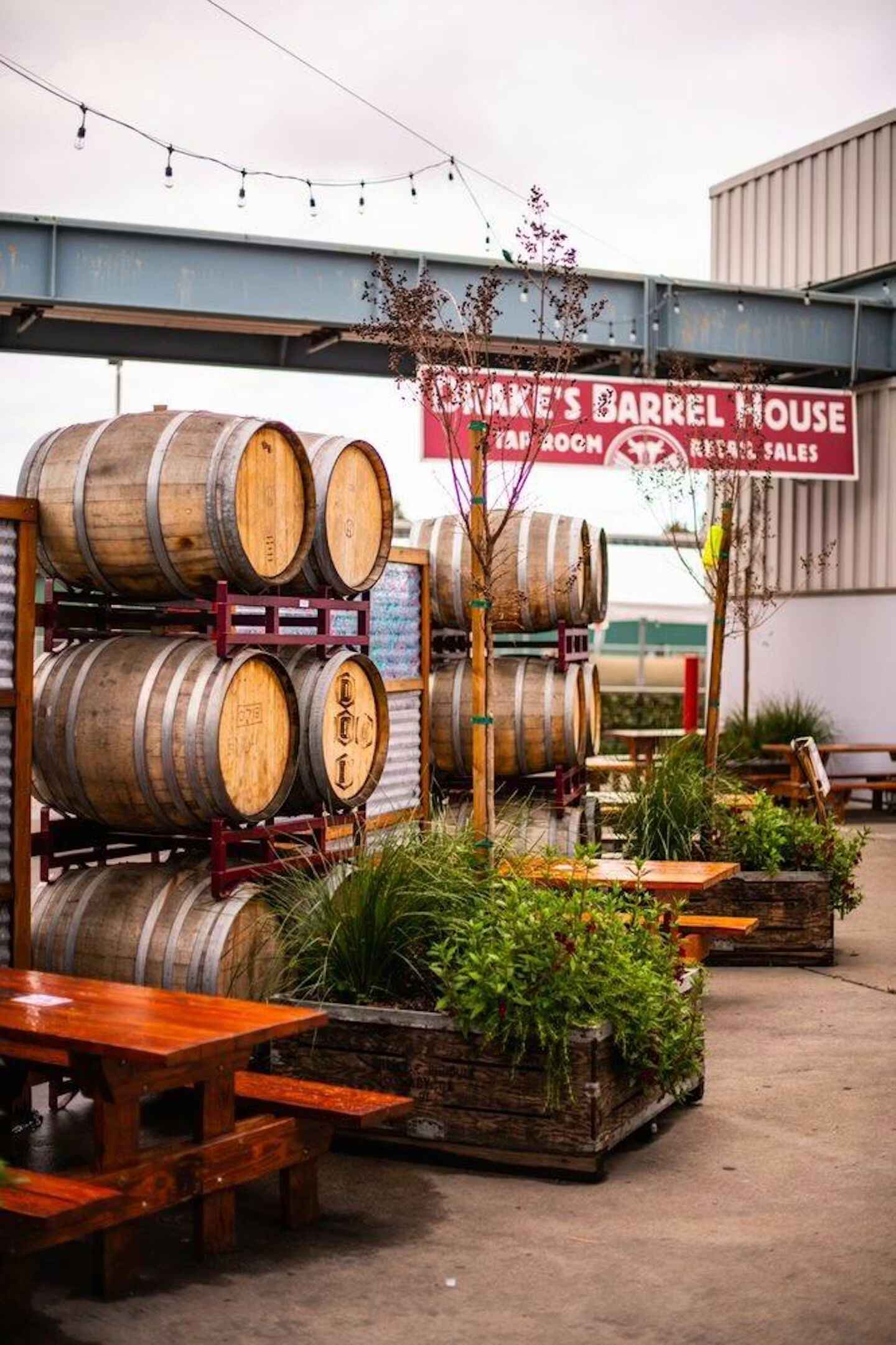 Outdoor seating area with wooden barrels stacked on racks, picnic tables, plants, and a red sign reading "Drake's Barrel House Tap Room & Retail Sales.