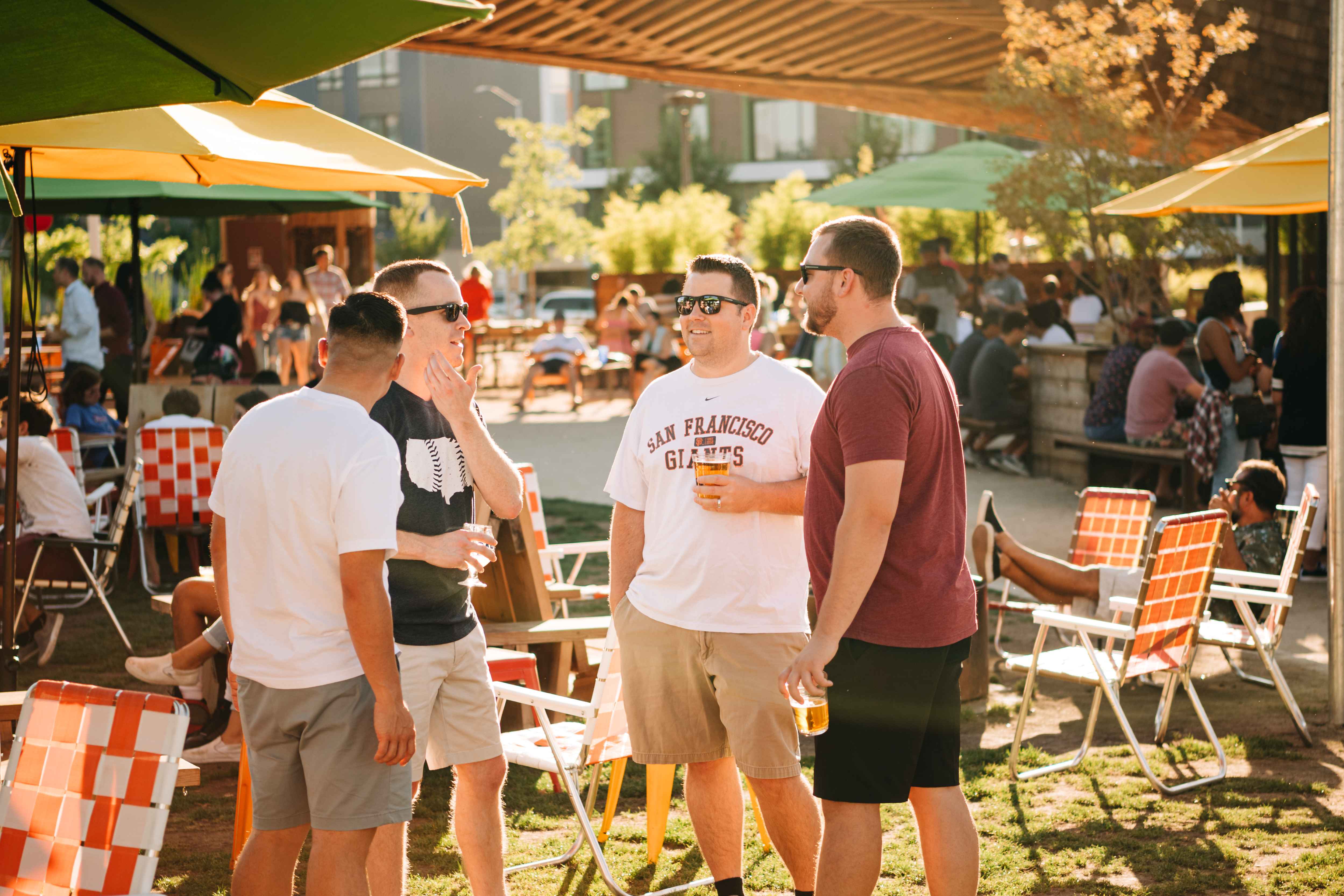 Four men in casual attire enjoy drinks at an outdoor patio bar on a sunny day