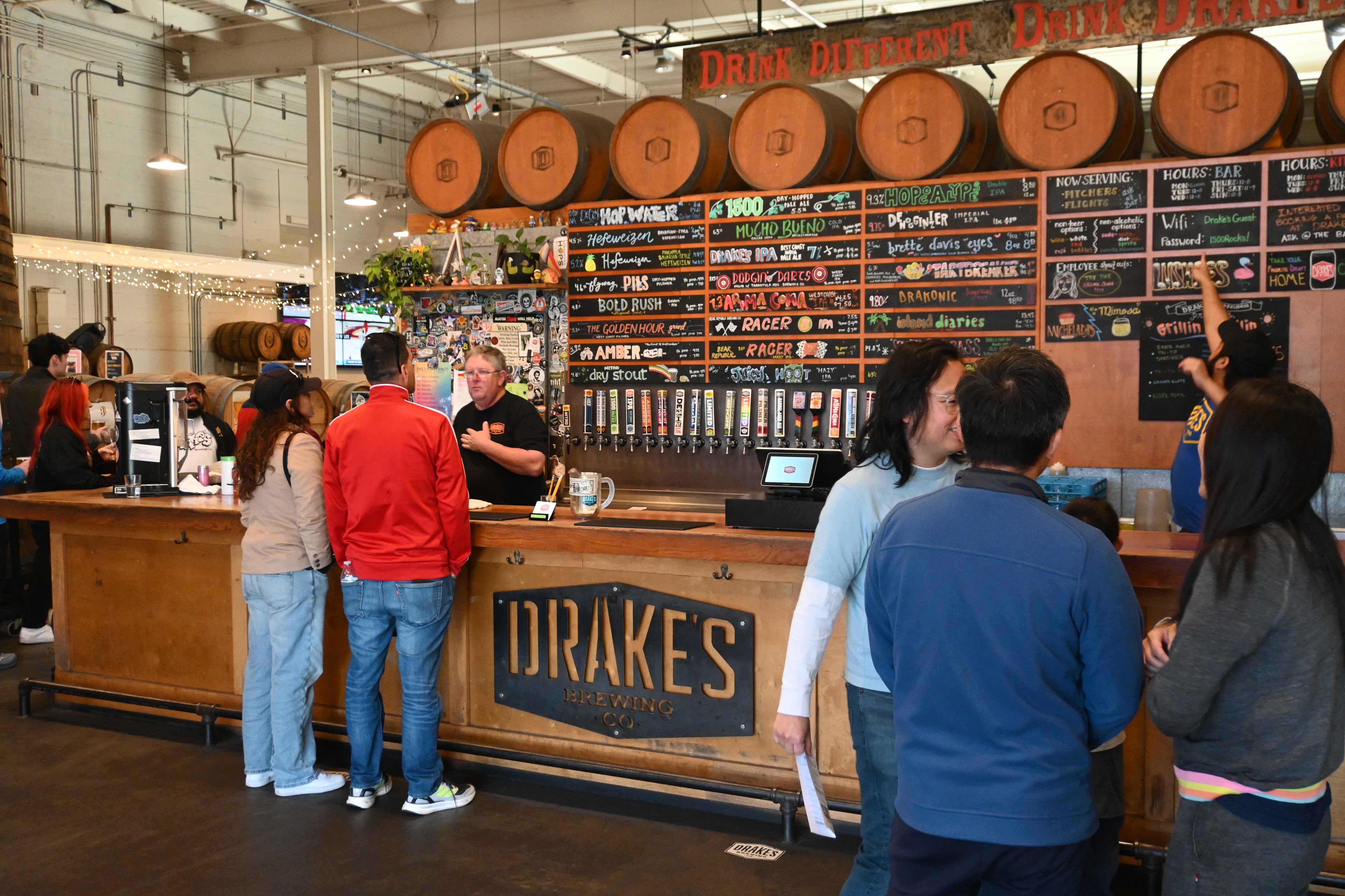 Customers order drinks at the bar of Drake's Brewing, with large barrels and a colorful menu board displayed behind the counter