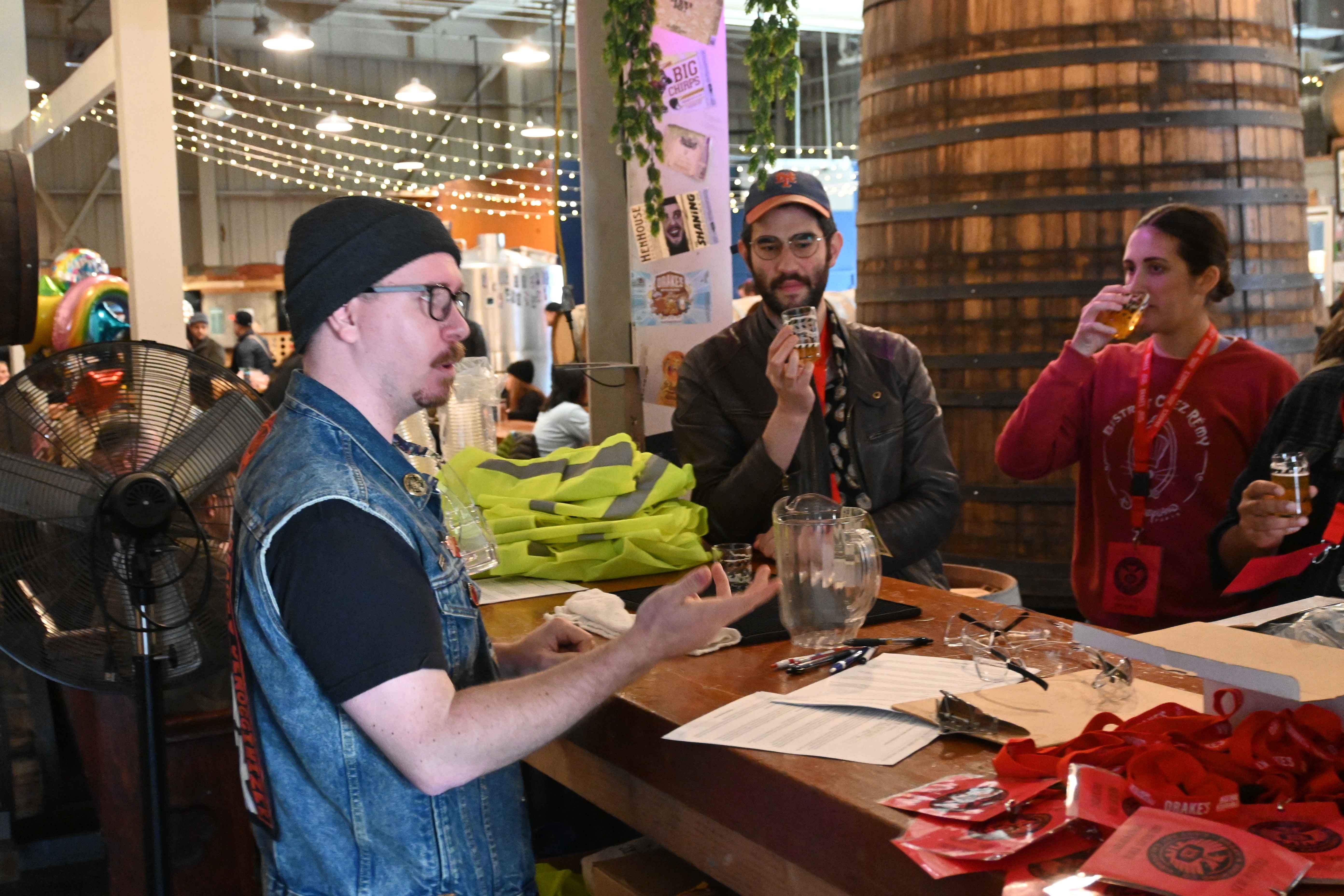 Three people sitting around a wooden table tasting beverages in a lively indoor market or brewery setting