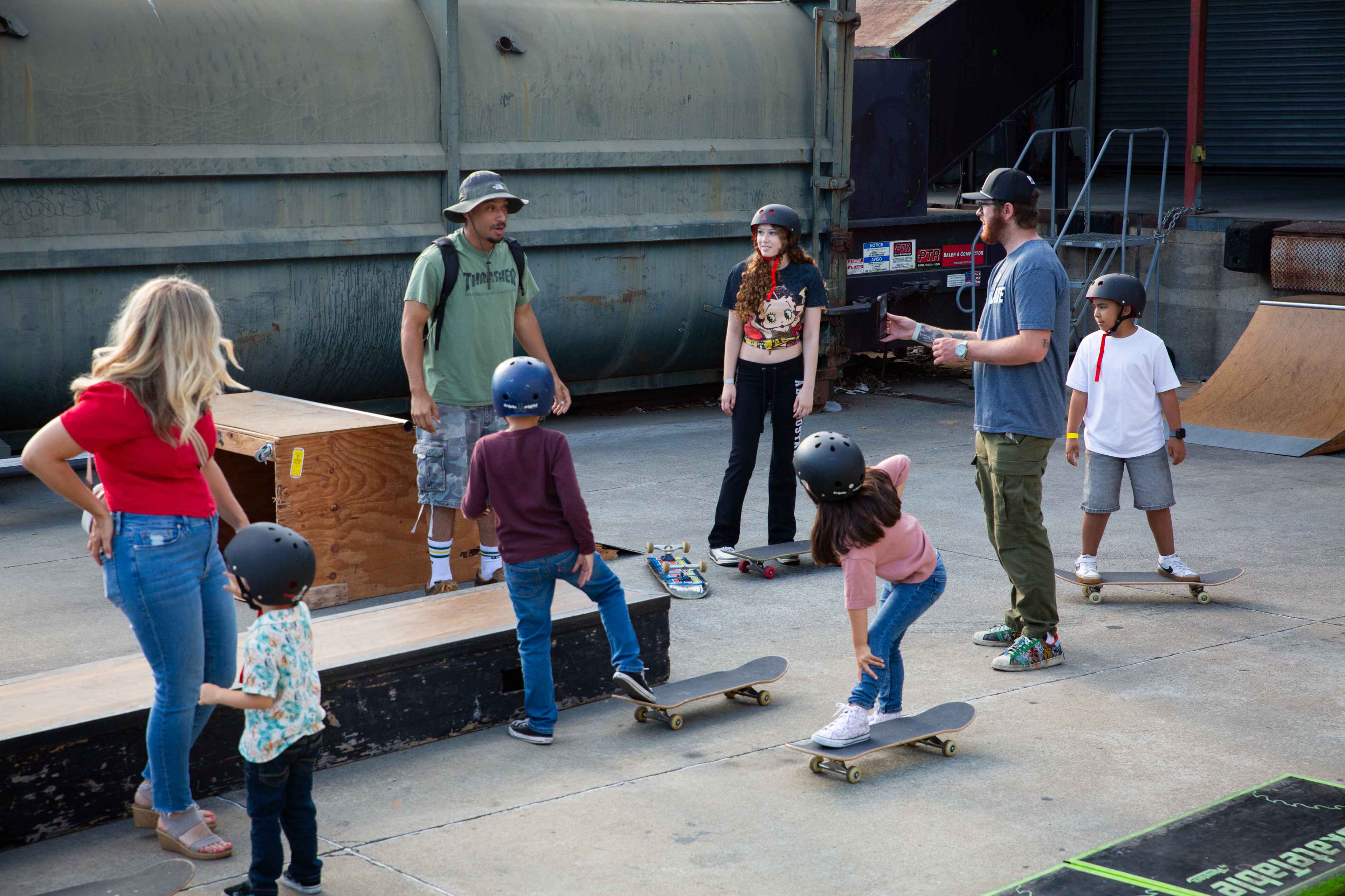 A group of children in helmets on skateboards listen to two instructors at an outdoor skate park while adults watch nearby