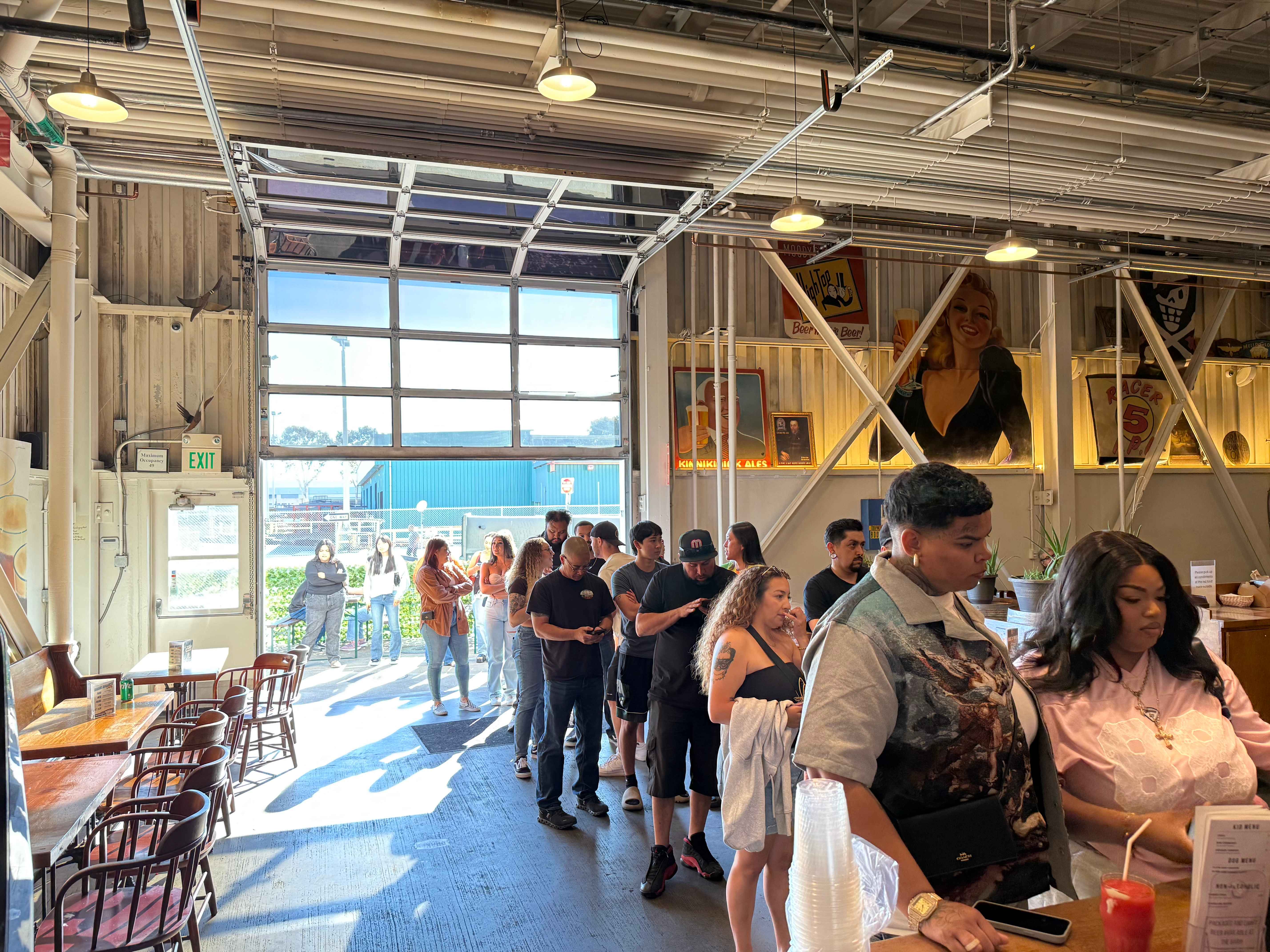 People wait in line inside a bright, spacious cafe with large windows and industrial decor