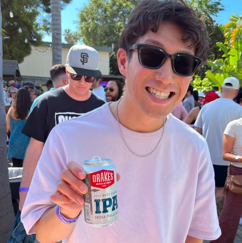 Man in sunglasses smiles, holding a Drakes IPA can at an outdoor event