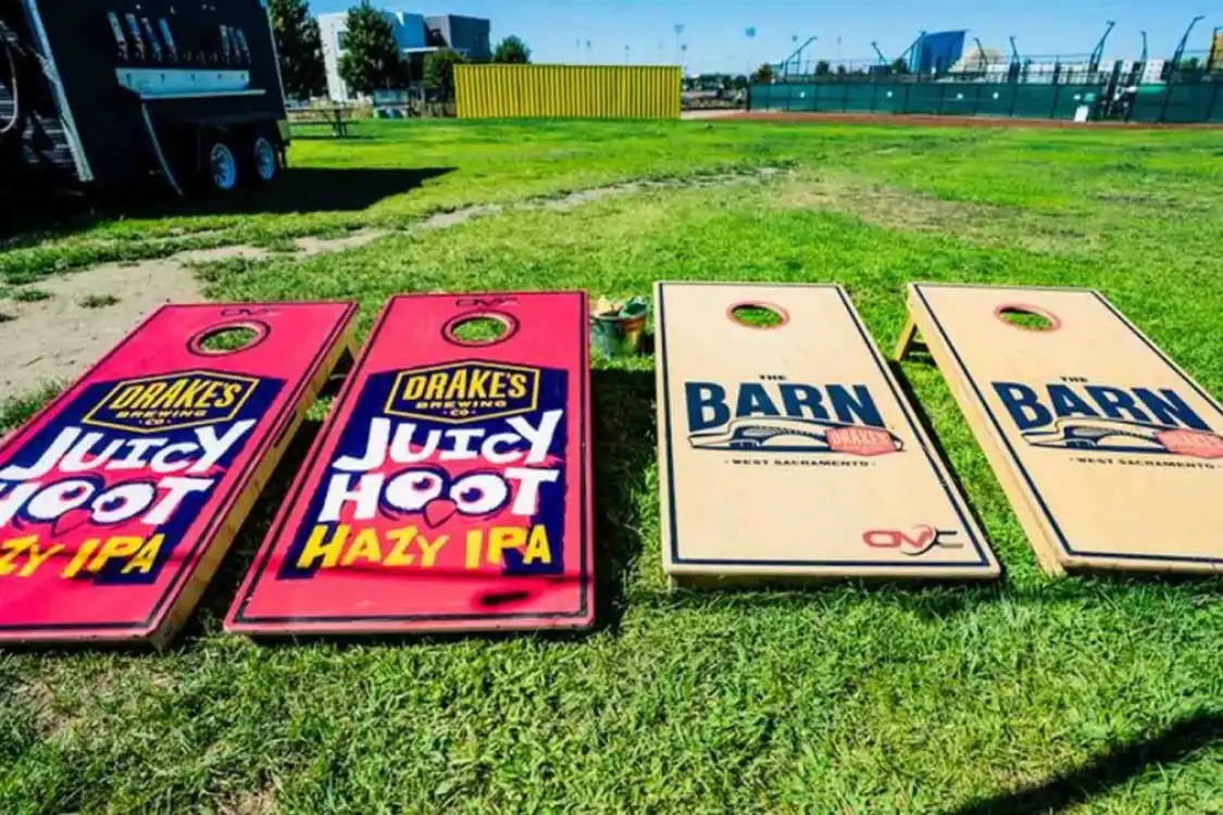Four cornhole boards on grass, two with "Drakes Juicy Hoot Hazy IPA" branding