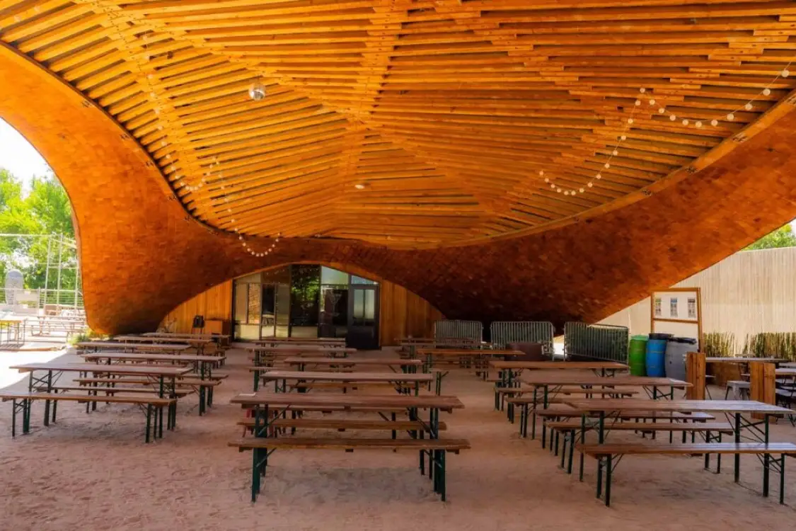Wooden picnic tables under a curved, slatted wooden roof, outdoor seating area